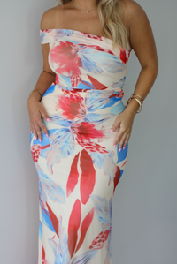 A woman modeling a strapless off-the-shoulder maxi dress with a vibrant red, blue, and cream floral print against a white wall. The photo shows a closeup of the bodice of the dress. 