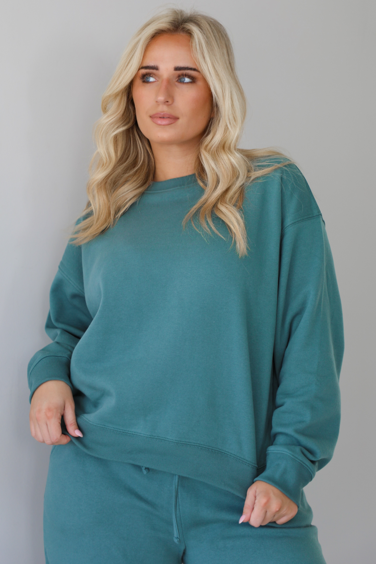 Woman wearing a sage two piece sweat-set. The set features a slightly oversized sweater and high waisted sweat pants that have an elastic and drawstring waistline with pockets. The sweater is untucked and shows the full length. The photo is taken indoors against a white wall with white marble flooring. 