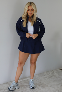 Woman wearing a navy tracksuit featuring a jacket and skort with a white tank indoors against a white wall with white marble flooring. 