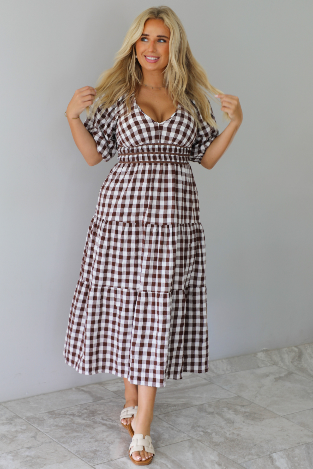 A woman modeling a brown and white gingham tiered midi dress with puff sleeves and a smocked waist, paired with beige slide sandals, standing indoors against a white wall with white marble flooring. 