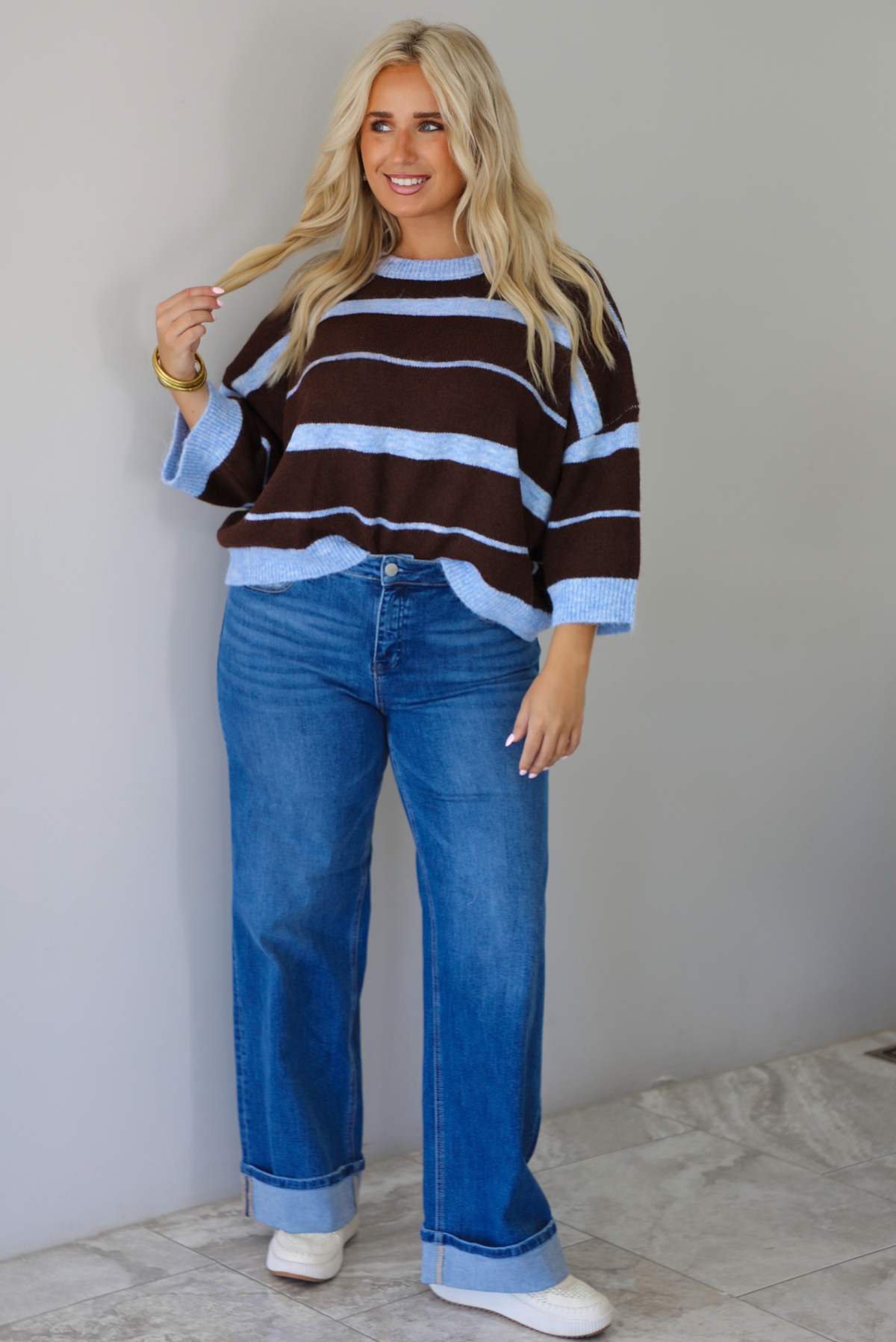 A woman wearing a brown and blue striped knit top with dark denim jeans, standing indoors against a white wall with white marble flooring. 