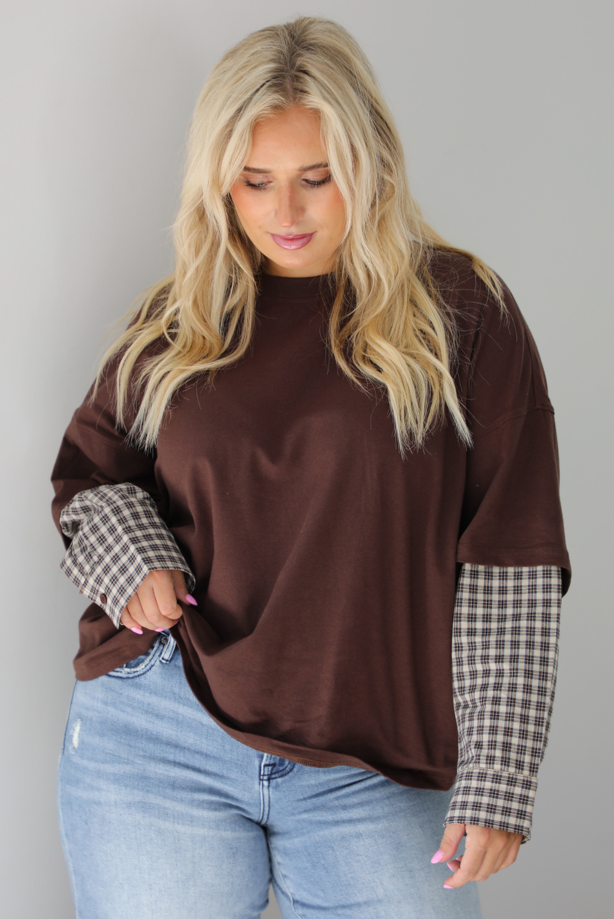 A woman wearing a brown t-shirt with a layered look of plaid sleeve detail, paired with light wash denim jeans, standing indoors against a white wall.