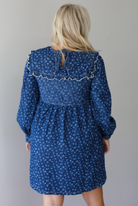 A woman shown from behind wearing a dark denim mini dress with a white floral print pattern throughout, collared neckline, a quilted detail at the bodice, long sleeves, and a flowy skirt, standing indoors against a white wall.The back shows a tie closure at the nape of the neck. 