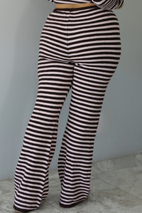 A woman wearing a light pink and brown striped two piece set featuring a double zipped hoodie top and mid-rise, wide leg pants, standing indoors against a white wall with white marble flooring. The photo shows a closeup of the back of the pants.