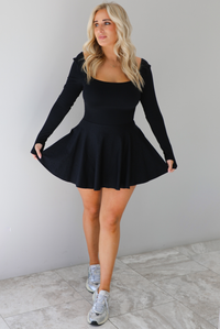 Woman wearing a black athleisure dress that has long sleeves with thumbhole cutouts, a scooped neckline, fitted bodice, and flowy skirt. The dress is paired with silver sneakers. The photo is taken indoors against a white wall with white marble flooring. 