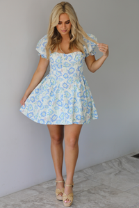 Woman wearing a white, blue, and green floral print mini dress that has short, puffed sleeves, a fitted corset bodice, and a flowy skirt indoors against a white wall with white marble flooring. 