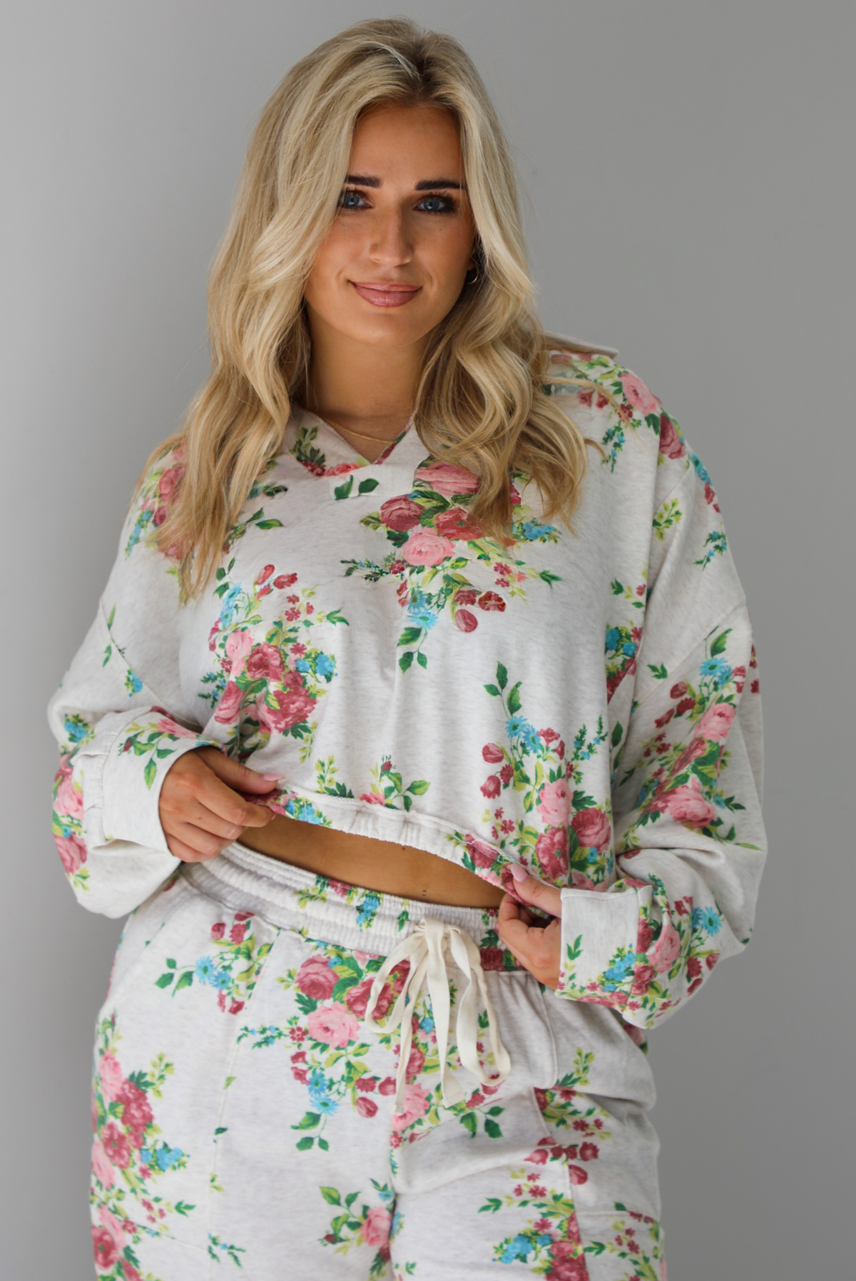 Woman wearing an oatmeal colored and multi-colored floral print two piece set featuring a collared long sleeved crop top and high waisted pants. The photo shows a closeup of the top. The photo is taken indoors against a white wall.