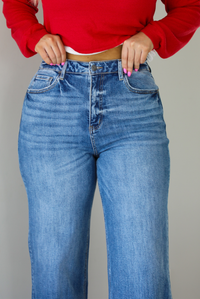 Woman wearing a pair of medium wash denim that are high waisted, have functional button and zipper closures, functional front and back pockets, a straight leg design, and a raw, distressed hem. The photo shows a closeup of the waistline of the jeans. The photo is taken indoors against a white wall.