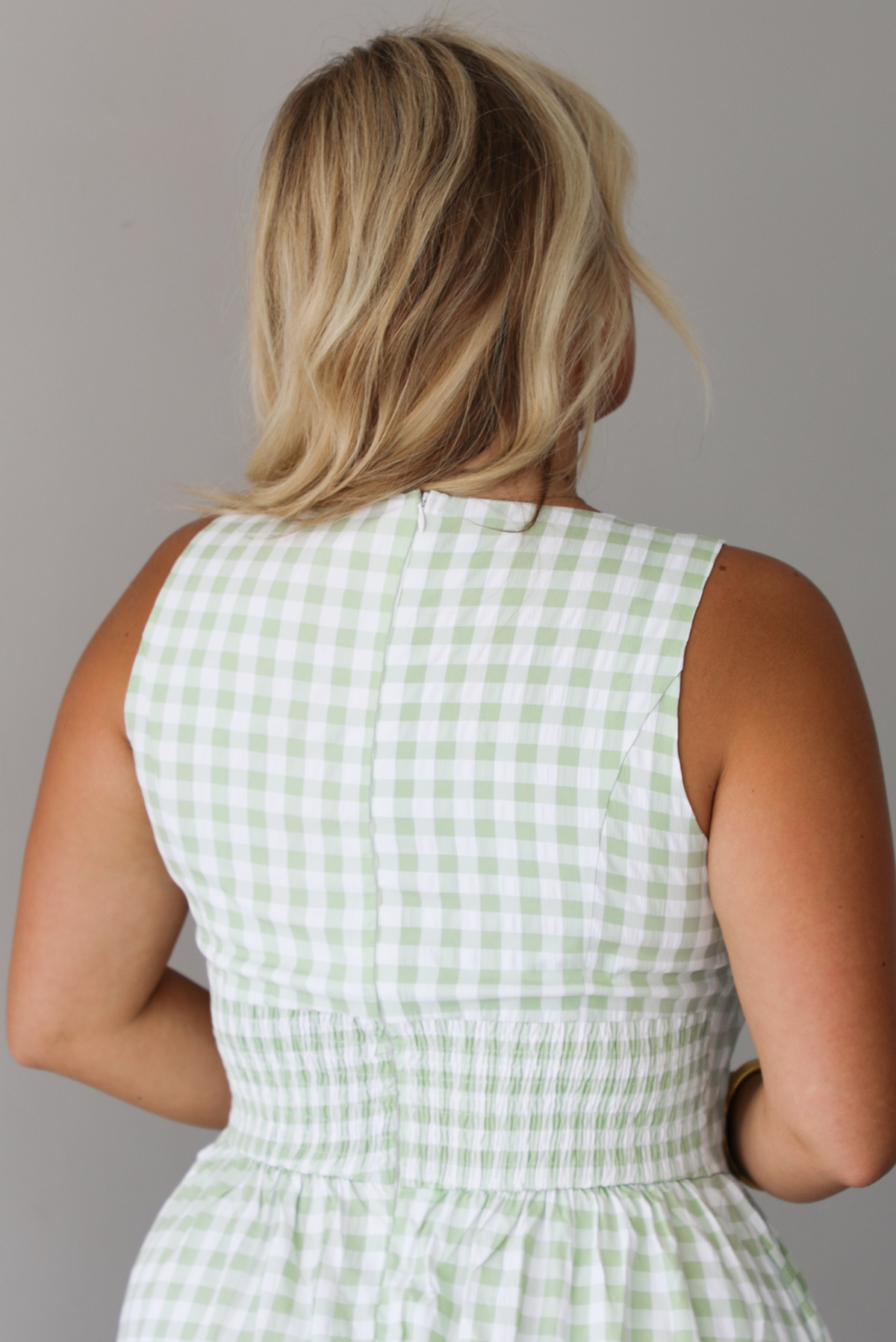 A woman shown from behind wearing a light green and white gingham maxi dress with a drop waist bodice and side pockets, standing indoors against a white wall. The back is shown to have a zipper closure and smocked waistline.
