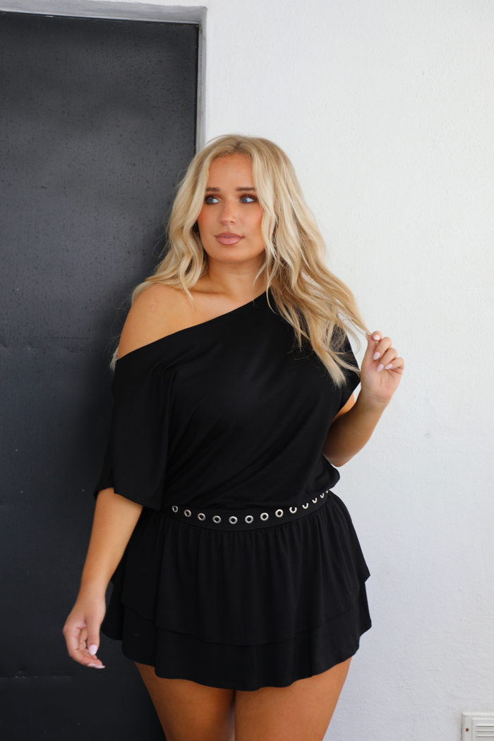 Woman model wearing a black dress that has a loose fit top with a cinched detail that sits at the hips and has silver circle studded details, and a ruffled skirt. The sleeve of the dress is worn off one shoulder.  The photo is taken outdoors against a white wall with a black door in the back.