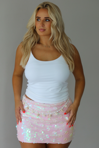 A blonde woman posing in a white ribbed tank top and a light pink sequined mini skirt with a beaded belt, indoors against a white wall. 