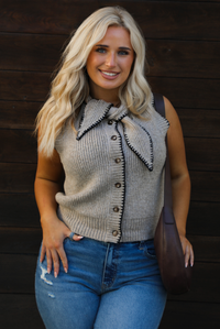 Close up of a woman model wearing a taupe sweater vest with black stitched detailing, has a tie detail at the neckline, is sleeveless, has functional button closures in the front, and has a ribbed texture. The sweater vest is paired with a medium wash denim, a dark brown leather purse, and ugg boots. The photo is taken outdoors against a dark brown wooden door. 