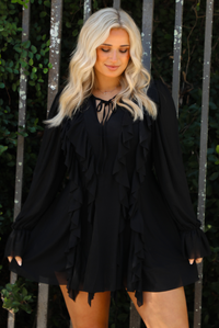 Woman model wearing a black romper that looks like a dress. The romper has a tie closure at the front with ruffle details along the front and long, billowy sleeves. The photo is taken outdoors against a metal gate and greenery. 