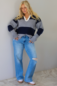 A woman wearing a navy and charcoal boyfriend style pullover top with a white collared detail, with distressed denim, standing indoors against a white wall with white marble flooring. 