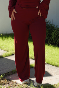 Close up of the pants of a woman model showcasing the bottoms of a two-piece burgundy wine colored set. The pants have a mid-rise, are fitted on the thighs, and lead to a straight leg design. They are lightweight, soft, and stretchy. The photo is taken outdoors against a white backdrop with grass at the bottom. The model is standing on cement pavers. 