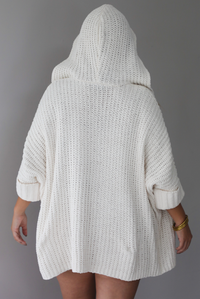 A woman shown from behind wearing a cream colored oversized cardigan with the hoodie up, standing indoors against a white wall.