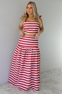 Woman wearing a red and white striped two piece set featuring a cropped top and maxi length, high waisted skirt. The photo is taken indoors against a white wall with white marble flooring. 