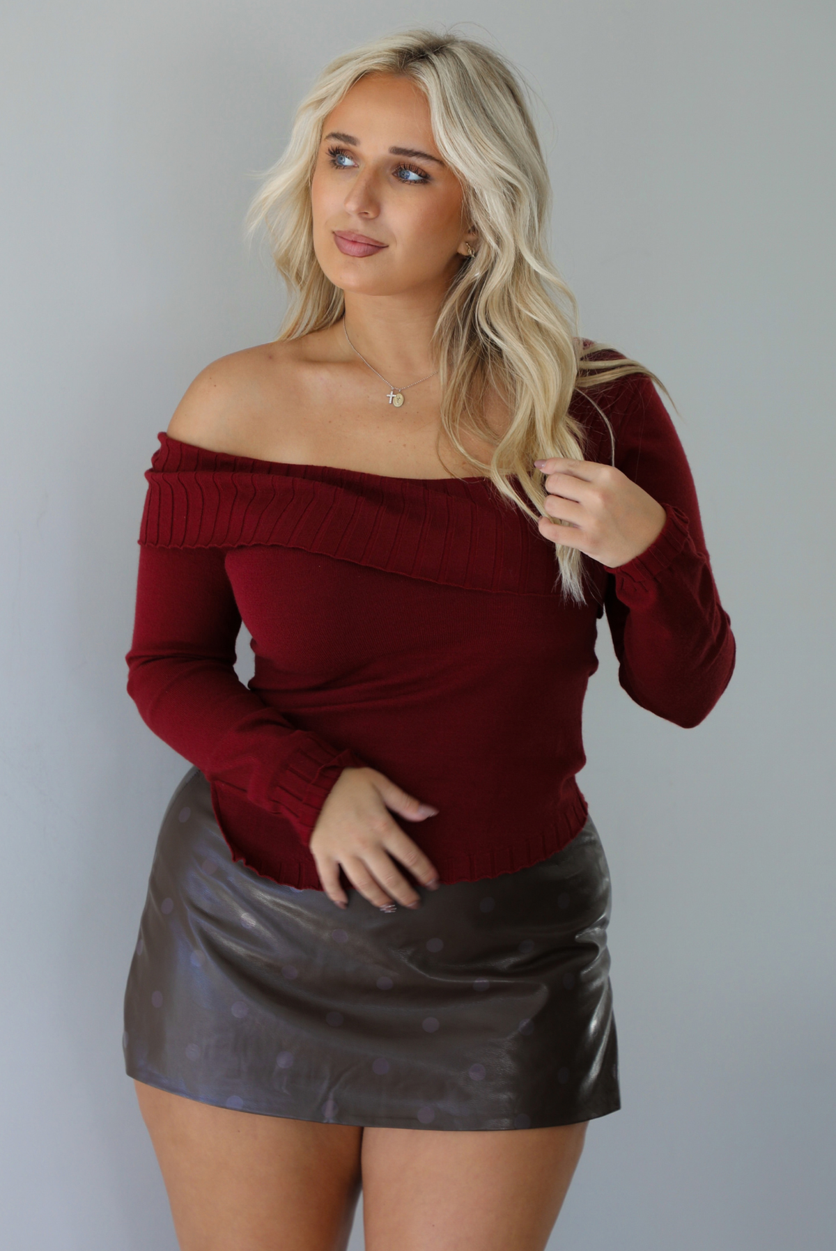Woman wearing a maroon knit, long sleeved, off the shoulder knit top paired with a dark brown vegan leather mini skirt with a monochromatic polka dot print pattern, standing indoors against a white wall.