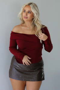 Woman wearing a maroon knit, long sleeved, off the shoulder knit top paired with a dark brown vegan leather mini skirt with a monochromatic polka dot print pattern, standing indoors against a white wall.