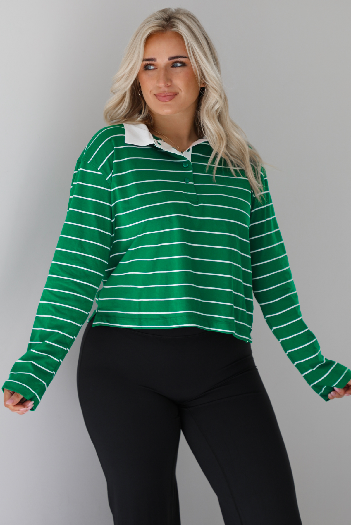 Woman wearing a green with white striped collared cropped top with functional button closures a quarter at the top. The top is paired with black leggings. The photo is taken in front of a white wall.
