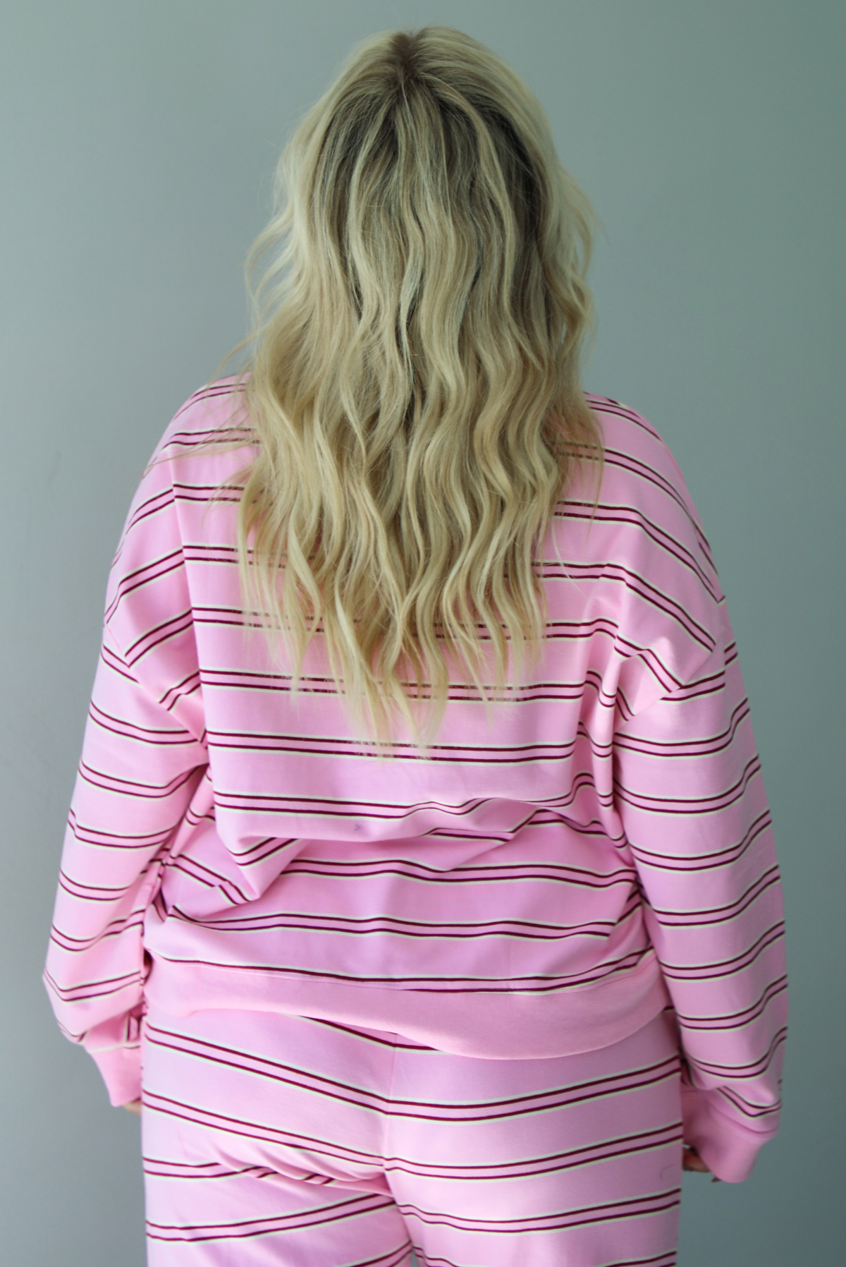 A woman shown from behind wearing a pink two piece sweat set featuring a maroon and white striped design on a sweatshirt and high waisted, wide leg pants, standing indoors against a white wall. The photo shows a closeup of the sweatshirt, which has a thick ribbed neckline and the same thick ribbed material at the wrists and bottom hem. 