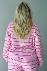 A woman shown from behind wearing a pink two piece sweat set featuring a maroon and white striped design on a sweatshirt and high waisted, wide leg pants, standing indoors against a white wall. The photo shows a closeup of the sweatshirt, which has a thick ribbed neckline and the same thick ribbed material at the wrists and bottom hem. 