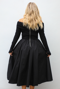 Woman wearing a black off the shoulder, long sleeved midi length dress with a pair of clear strapped nude heels. The photo shows the back of the dress, which is similar to the front except it also has a zipper closure. The photo is taken outdoors against a white wall.