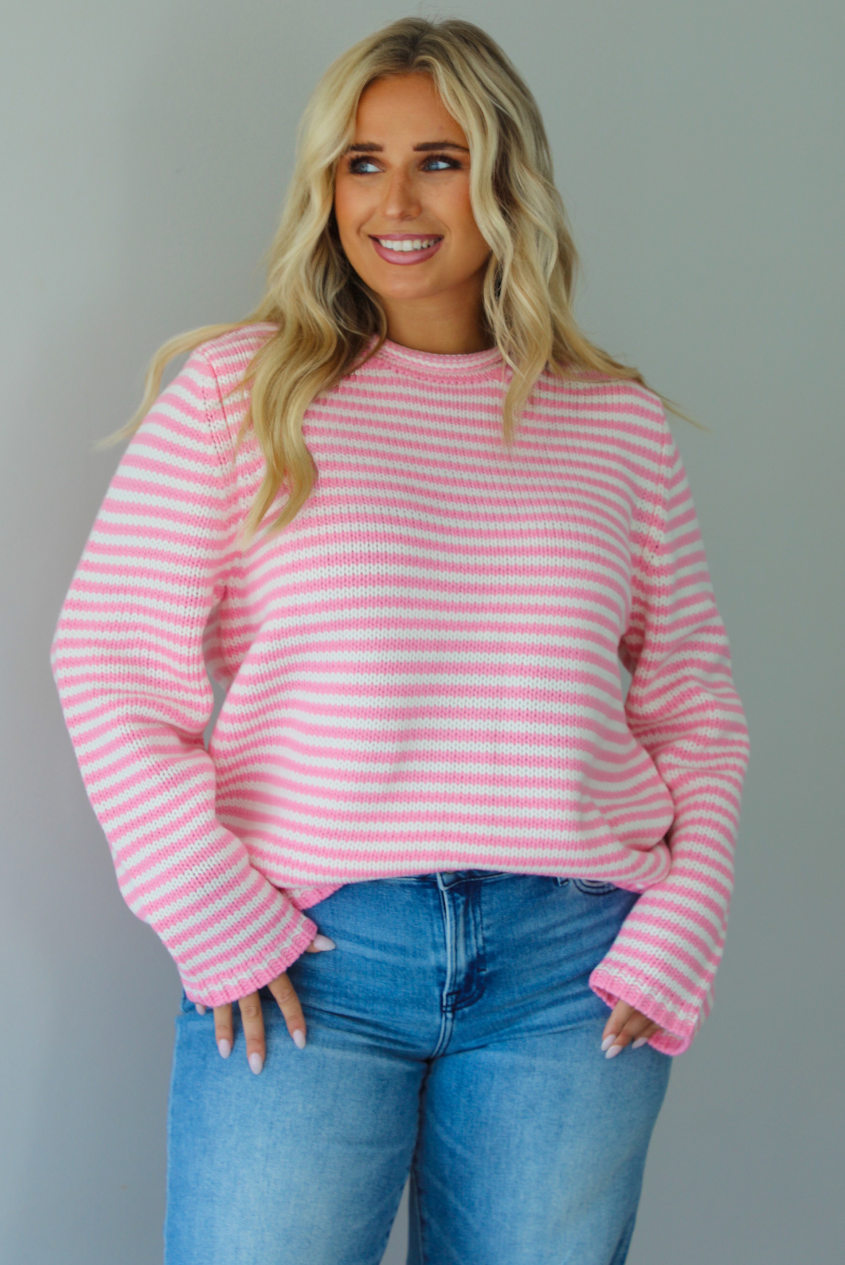 A woman wearing a pink and white striped sweater paired with light wash denim jeans, standing indoors against a white wall.