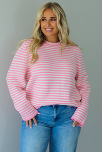 A woman wearing a pink and white striped sweater paired with light wash denim jeans, standing indoors against a white wall.