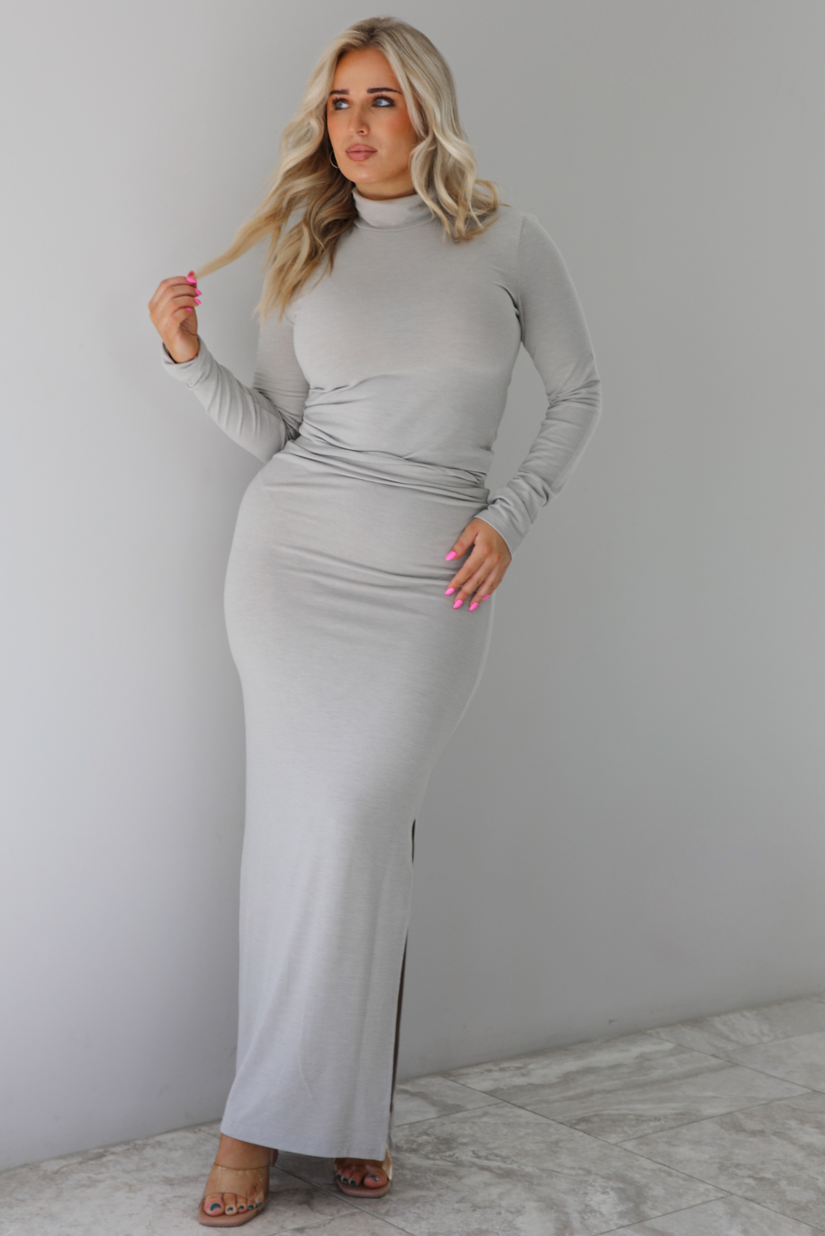 Woman wearing a grey two piece set featuring a turtle neck long sleeved fitted top and high waisted maxi length fitted skirt. The set is paired with clear strapped heels. The photo is taken indoors against a white wall with white marble flooring. 