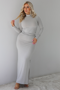 Woman wearing a grey two piece set featuring a turtle neck long sleeved fitted top and high waisted maxi length fitted skirt. The set is paired with clear strapped heels. The photo is taken indoors against a white wall with white marble flooring. 