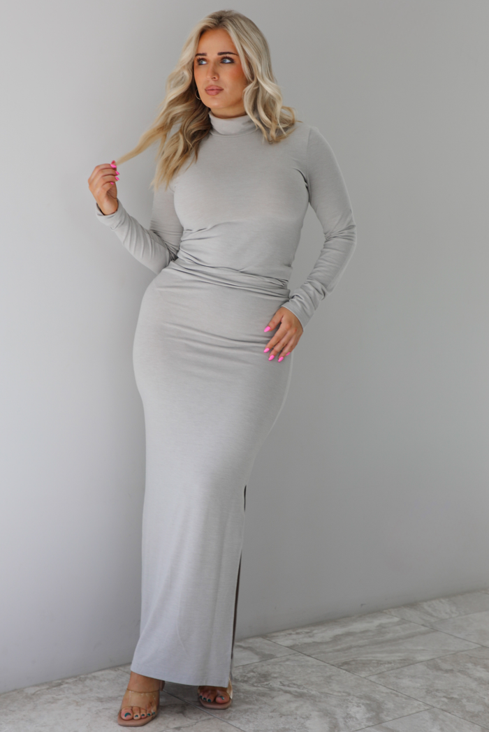 Woman wearing a grey two piece set featuring a turtle neck long sleeved fitted top and high waisted maxi length fitted skirt. The set is paired with clear strapped heels. The photo is taken indoors against a white wall with white marble flooring. 