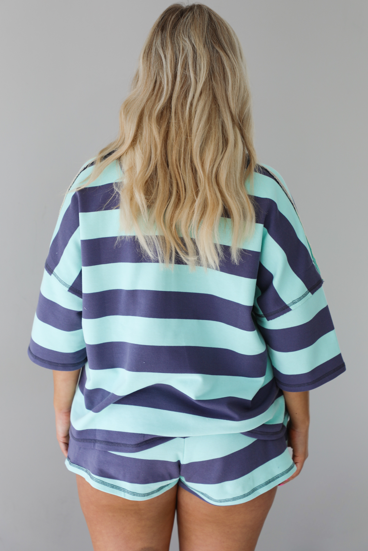 Woman wearing a turquoise and navy thick striped set featuring a collared short sleeved quarter zip top and high waisted shorts that have an elastic drawstring waistline with functional pockets on either side. The photo is taken indoors against a white wall. The photo shows a closeup of back of the set.