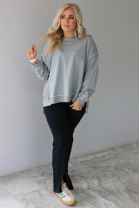 Woman wearing a grey oversized sweater with black leggings that have a slightly flared hem with split hem details in the front. The photo is taken in front of a white wall with white marble flooring. 