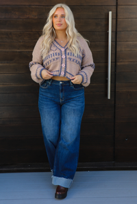Woman model wearing a mocha brown cardigan that has grey stitching details throughout. The cardigan has a cropped cut, is oversized, and has functional button closures in the front. She's wearing the cardigan with dark denim jeans and brown shoes. The photo is taken against a dark brown wooden door outdoors. 
