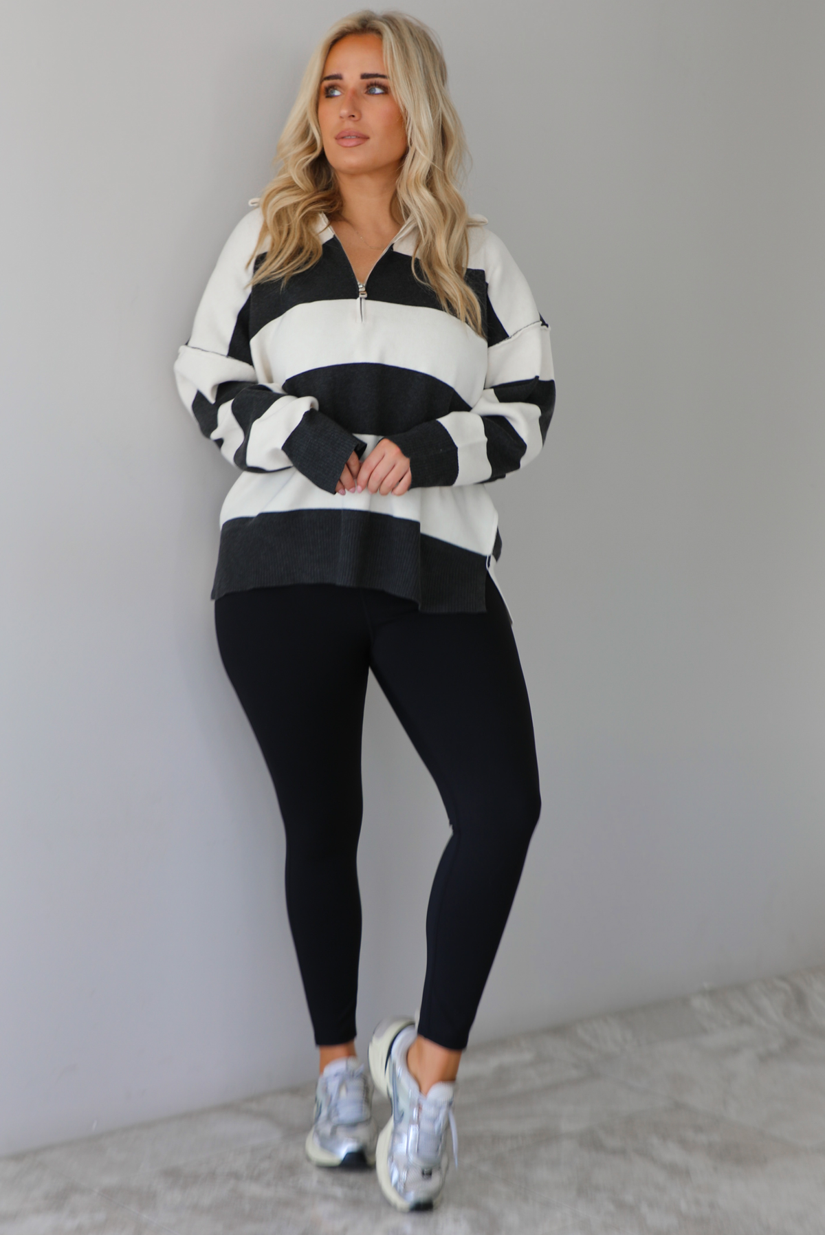 Woman wearing a charcoal and white striped quarter-zip sweater with a collared neckline, raw seam details, and a relaxed fit. It's paired with a fitted black jumpsuit and silver sneakers. The photo is taken indoors against a white wall with white marble flooring.