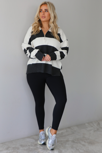 Woman wearing a charcoal and white striped quarter-zip sweater with a collared neckline, raw seam details, and a relaxed fit. It's paired with a fitted black jumpsuit and silver sneakers. The photo is taken indoors against a white wall with white marble flooring.