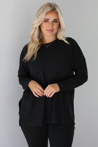 Woman wearing a black two piece set that features an oversized long sleeved top and fitted leggings. The photo shows a closeup of the top. The photo is taken indoors against a white wall.