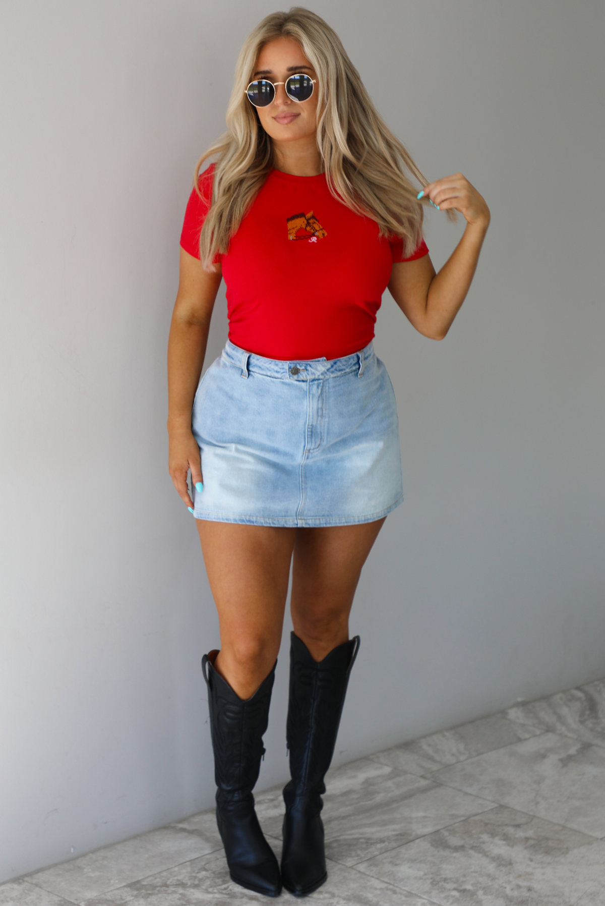 Woman wearing a red fitted baby tee with an embroidered horse graphic on the front. The top is paired with a high waisted, light-wash denim skort. The photo is taken indoors against a white wall with white marble flooring. 