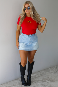 Woman wearing a red fitted baby tee with an embroidered horse graphic on the front. The top is paired with a high waisted, light-wash denim skort. The photo is taken indoors against a white wall with white marble flooring. 