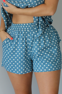 Woman wearing denim blue with white polka dot halter tie top and high waisted shorts indoors against a white wall. The photo shows a closeup of the shorts, which have an elastic waistline and functional pockets on either side. 