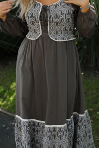 Close up of a woman model wearing an olive and off-white stitched detail dress outdoors. Dress has a corset-appearance bodice, long sleeves, functional pockets on either side, an elastic waistline, and a long, maxi length.