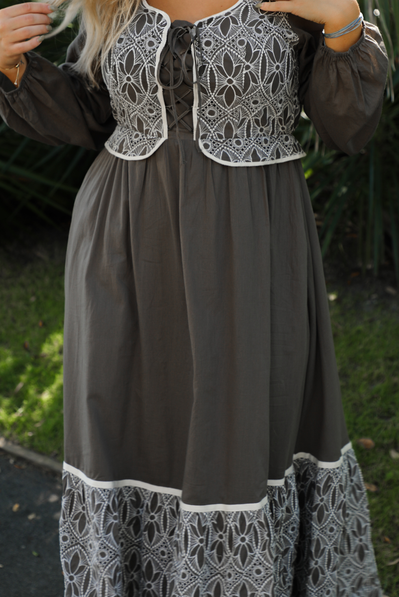 Close up of a woman model wearing an olive and off-white stitched detail dress outdoors. Dress has a corset-appearance bodice, long sleeves, functional pockets on either side, an elastic waistline, and a long, maxi length.