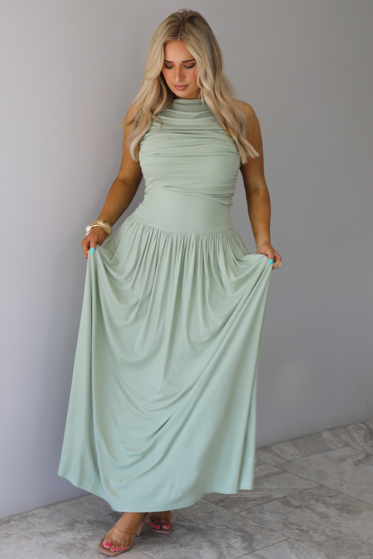 Woman wearing a sage green maxi length dress, which has a rouched detail at the bust, a fitted waistline, a flowy skirt. The photo is taken indoors against a white wall with white marble flooring. 