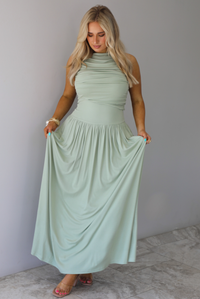 Woman wearing a sage green maxi length dress, which has a rouched detail at the bust, a fitted waistline, a flowy skirt. The photo is taken indoors against a white wall with white marble flooring. 