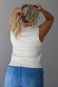A woman shown from behind wearing a cream colored sweater vest with a mallard duck on the front, paired with medium wash denim with wide leg hem, standing indoors against a white wall.