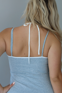 Woman wearing a heather grey athleisure dress with white trim detailing at the hem indoors against a white wall. The photo shows the back of the dress, along with the adjustable straps, and the halter tie neckline. 