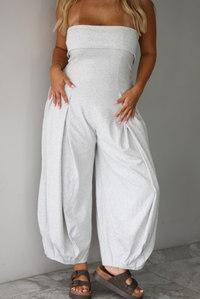 A woman modeling a light gray strapless jumpsuit with wide, balloon-style legs and gathered detailing, standing indoors against a white wall with white marble flooring. 