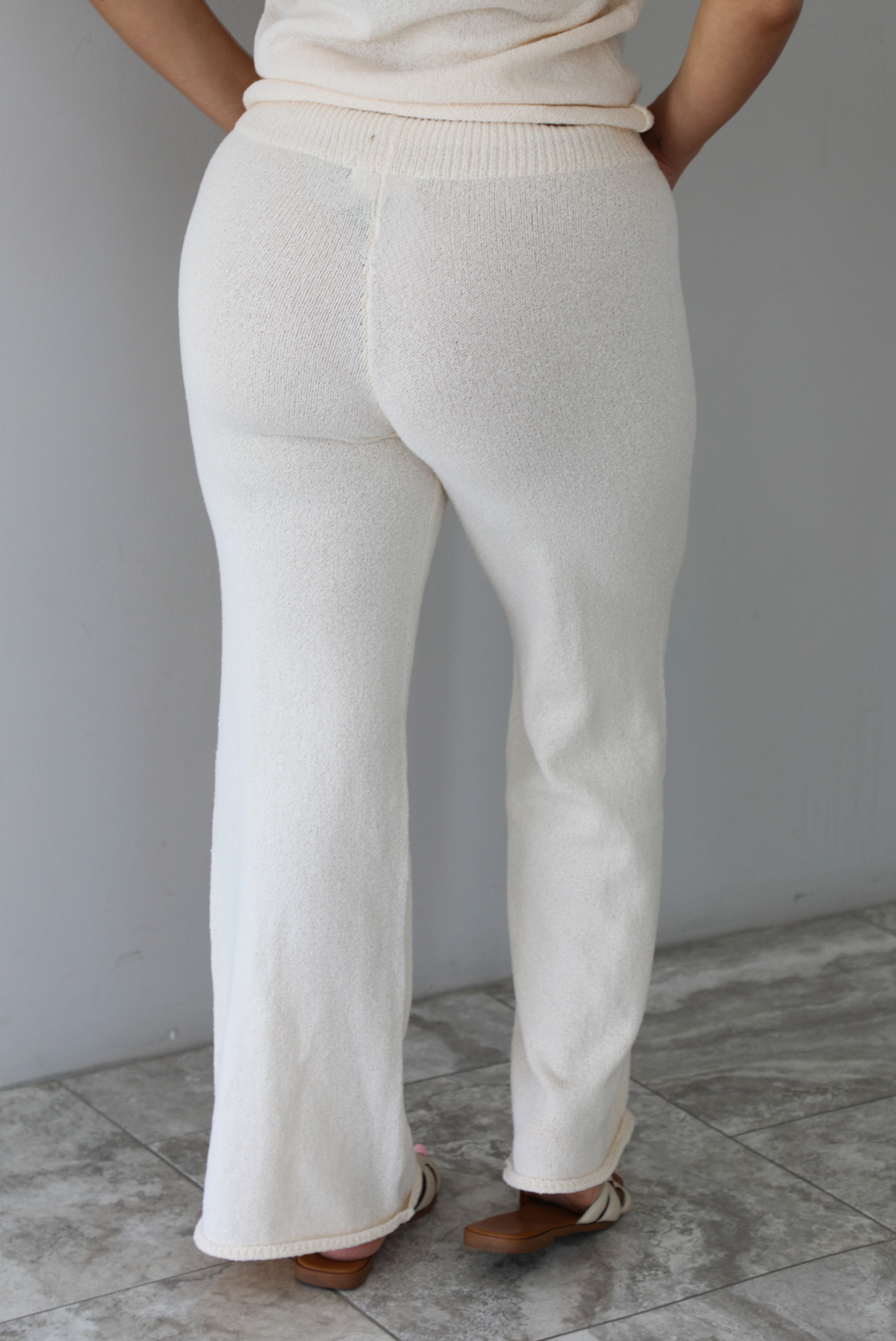 Woman wearing a cream two piece set featuring a high-neck sleeveless top and matching mid-rise pants indoors against a white wall with white marble flooring. The photo shows a closeup of the back of the pants.