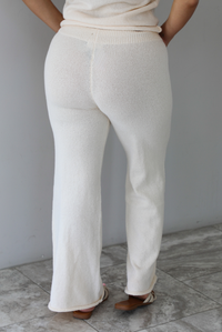 Woman wearing a cream two piece set featuring a high-neck sleeveless top and matching mid-rise pants indoors against a white wall with white marble flooring. The photo shows a closeup of the back of the pants.